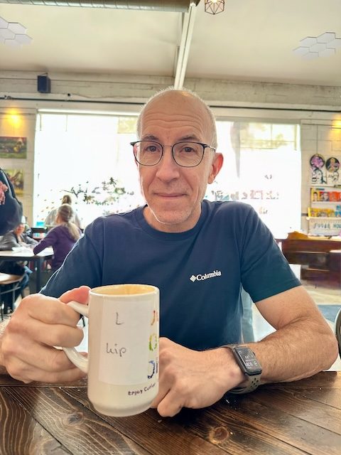Picture of man holding white mug seated at a table in front of some windows at Sugarhouse Coffee/