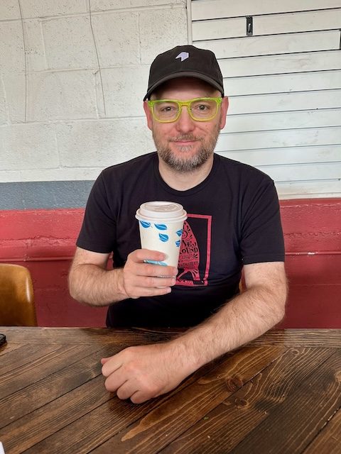 Man with black shirt sitting against the wall with a paper cup holding a hot drink.